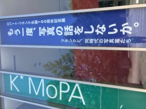 外観バナー。正面入り口側、ガラス張りの吹き抜けに、縦に細長いバナーが「K*MoPA」と今回の企画展の名前の計2本掛かっている様子。